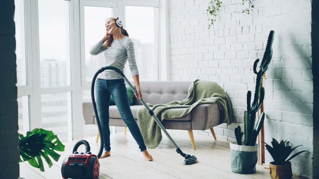Beautiful young woman is hoovering the floor at home using modern vacuum cleaner and listening to music with headphones, dancing and singing. Housework and technology concept.