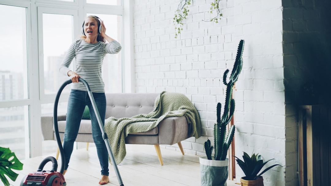 Cheerful blond housewife is using vacuum cleaner during routine clean-up at home, listening to music through headphones, singing and dancing. Technology and young people concept.